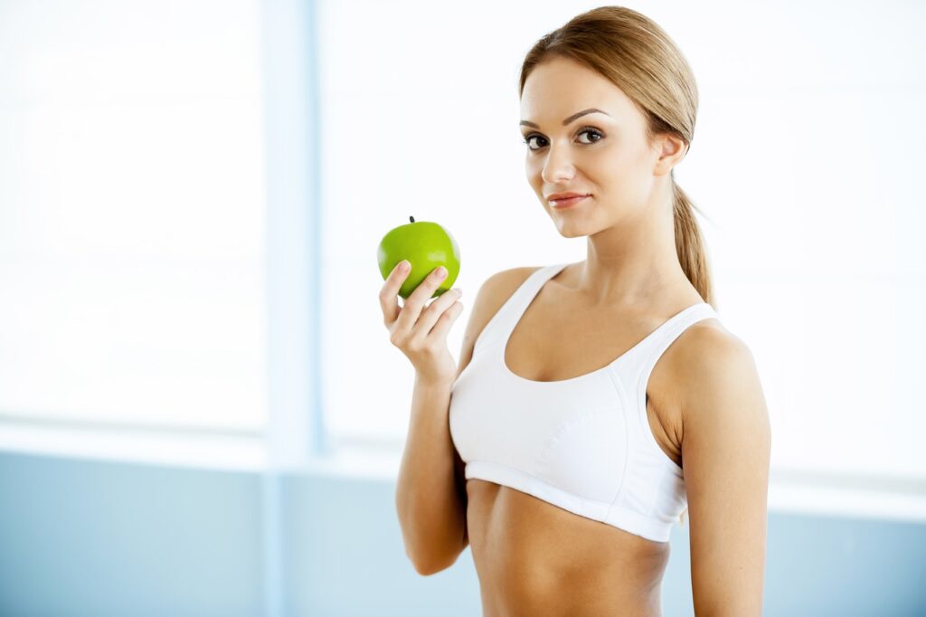 Sport and dieting. Beautiful young woman in sports clothing holding green apple and smiling at camera